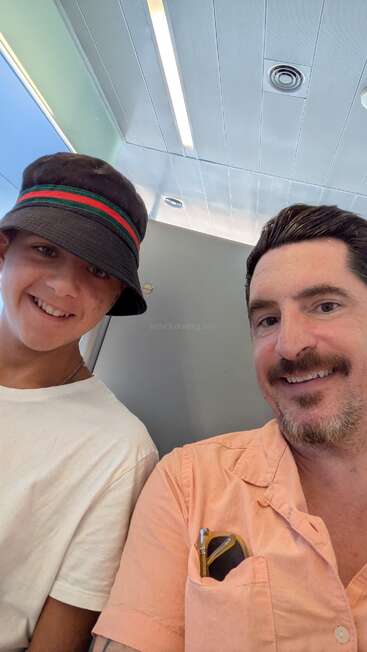 Ein lächelnder Mann in einem pfirsichfarbenen Hemd und ein Junge mit Eimerhut und weißem T-Shirt machen zusammen ein Selfie in einem hell erleuchteten Innenraum.