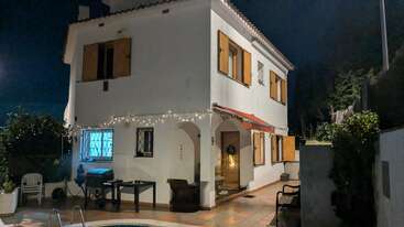 Ein zweistöckiges weißes Haus bei Nacht, mit hölzernen Fensterläden, Außenbeleuchtung, einer Terrasse, Stühlen, einem Tisch und einem Swimmingpool, umgeben von Bäumen und Büschen.