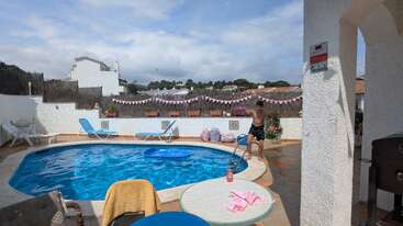 Eine sonnige Szene am Pool mit Liegestühlen, Handtüchern und Dekorationen. Ein Mann steht am Wasser. Helle, entspannte Atmosphäre mit Hügeln und Häusern im Hintergrund.