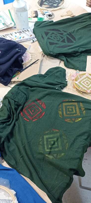 Two green shirts with stenciled geometric designs lie on a messy table, surrounded by paints, paintbrushes, cardboard, and art supplies, showcasing a creative workspace.