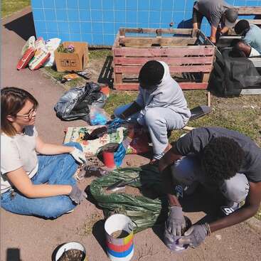 Un groupe de personnes jardine ensemble, remplissant des récipients de terre et travaillant près de caisses en bois. Ils semblent concentrés, collaborant en plein air sous un soleil radieux.