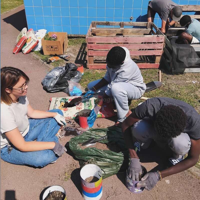 A group of people is gardening together, filling containers with soil, and working near wooden crates. They appear focused, collaborating outdoors under bright sunlight.