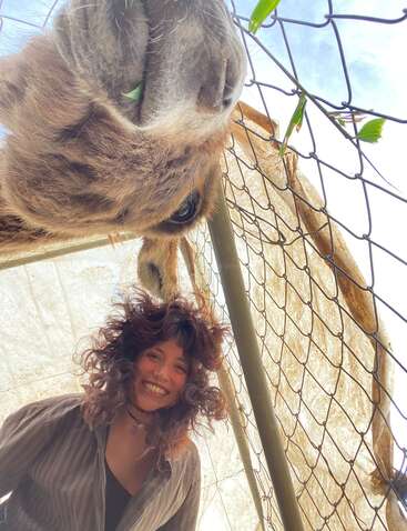 Une femme rayonnante aux cheveux bouclés sourit sous le regard curieux d'une girafe photographiée en gros plan derrière une clôture grillagée ; toutes deux regardent vers l'appareil photo par une journée ensoleillée.