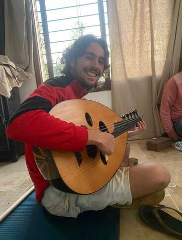 Un homme souriant, vêtu d'une veste rouge et noire, est assis par terre et joue de l'oud. La lumière naturelle qui pénètre par la fenêtre derrière lui crée une atmosphère chaleureuse.