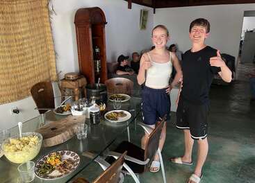 Deux personnes souriantes se tiennent près d'une table à manger en verre garnie de plats et lèvent le pouce. D'autres se détendent à l'arrière-plan. On dirait un repas convivial et décontracté entre amis.