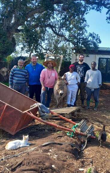 Six people stand together outdoors, smiling by a donkey. There’s farming equipment, a cat, and a duck nearby. Trees and a small building appear in the background.