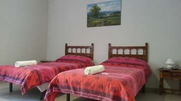 A imagem mostra um quarto com duas camas, cada uma com uma estrutura branca e um colchão cinza, acompanhadas de uma mesa de cabeceira e uma cômoda, todas situadas em um espaço bem iluminado e organizado.