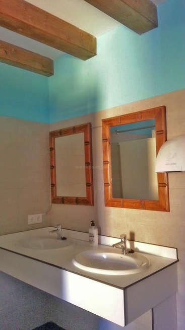 A imagem mostra um banheiro com uma pia branca, dois espelhos e uma luminária, com teto azul e vigas de madeira, contra uma parede bege.