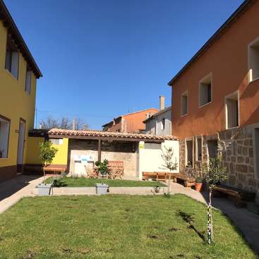 The image depicts a serene courtyard with a grassy area, surrounded by two-story buildings, featuring a bench, trees, and a covered seating area, set against a clear blue sky.