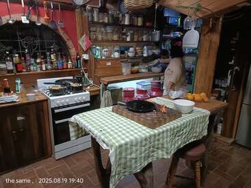 A rustic kitchen with wooden shelves, jars, and cooking supplies. A person stands at the counter. Table with green checkered cloth, cutting board, and oranges. Cozy atmosphere.