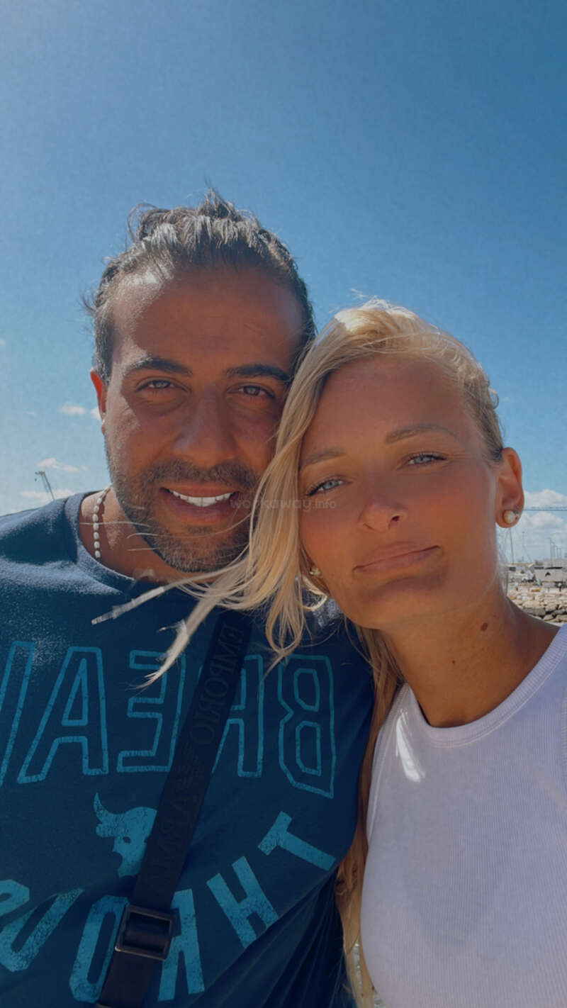 A smiling couple poses for a selfie outdoors on a sunny day. Blue sky, marina background, relaxed summer attire, and warm expressions capture a joyful moment together.