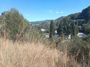 A imagem retrata uma paisagem montanhosa serena com uma estrada sinuosa, árvores e alguns edifícios, em um céu azul claro.