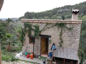 A imagem mostra uma casa de pedra rústica com porta e chaminé de madeira, cercada por uma vegetação exuberante, em um cenário natural sereno.