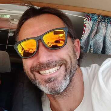 A imagem mostra um homem com cabelos escuros e barba, usando óculos escuros, no banco de trás de um carro com um painel de madeira acima da cabeça.