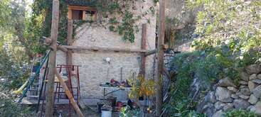 A imagem mostra um edifício rústico de pedra com uma janela de madeira, cercado por um jardim e equipamentos de construção, o que sugere um trabalho de renovação ou reparo em andamento.