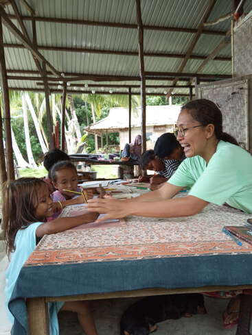 A smiling woman interacts joyfully with two young girls at a table, engaging in a fun activity. The setting is a rustic, open-air structure.