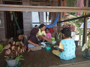 Four children are sitting together on a wooden porch, sharing a meal. Potted plants decorate the area, and hammocks hang near a cozy, wooden house.