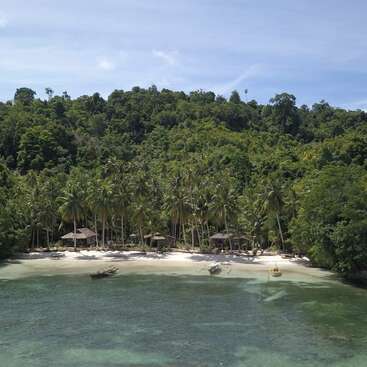 A secluded tropical beach with white sand, clear turquoise water, boats, palm trees, wooden huts, and lush green forest under a bright blue sky.