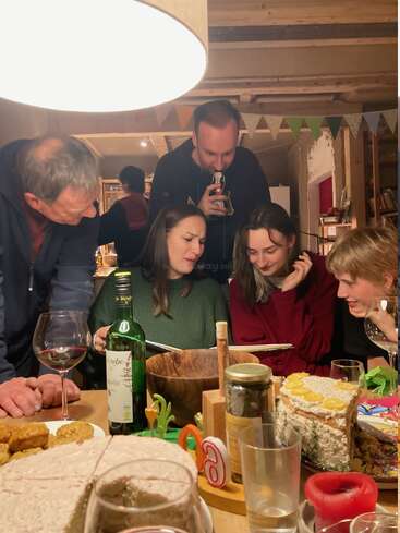 Cinco pessoas estão reunidas em torno de uma mesa cheia de vinho, bolo e petiscos, sorrindo e curtindo o momento enquanto observam algo juntas em uma sala aconchegante.