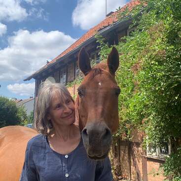 Uma mulher sorridente, de cabelos grisalhos, está ao lado de um cavalo castanho, ao ar livre num dia ensolarado, em frente a uma charmosa casa de tijolos coberta de vegetação.