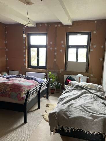 Esta imagem mostra um quarto simples e ainda sem acabamento, com duas camas de solteiro, paredes sem reboco, duas janelas, vasos de plantas, roupa de cama simples e uma única lâmpada pendurada.