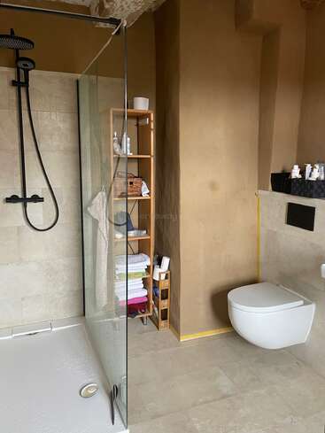 This bathroom features a modern walk-in shower with a black rainfall showerhead, a wooden shelf with towels and toiletries, and a wall-mounted toilet with minimalist decor.