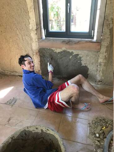 A smiling man sits on the floor by a window, applying plaster to a wall. He wears shorts, a blue shirt, and sandals, enjoying the renovation work.