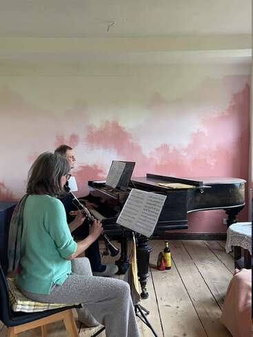 Two people are playing music in a cozy room. One plays clarinet, the other piano. Sheet music is on stands, and the walls are softly pink.