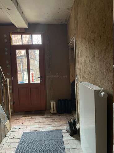 This image shows a rustic entryway with brick flooring, a wooden door, several pairs of boots, exposed beams, a radiator, and textured, unfinished walls.