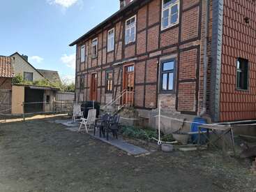 A brick and timber house with chairs and a table in the backyard, dirt ground, outbuildings, blue sky, and a residential suburban neighborhood setting.