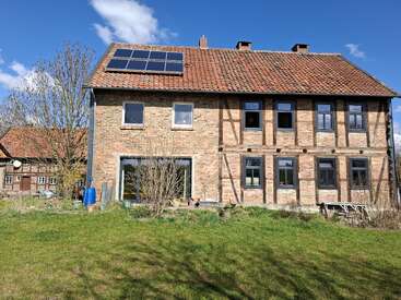 A rustic brick house with a red tiled roof and solar panels sits surrounded by grass, trees, and blue sky on a bright sunny day.