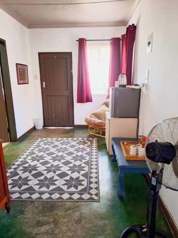 Une pièce chaleureuse avec un sol vert, un tapis à motifs, une porte marron, des rideaux rouges, des murs blancs, un mini-réfrigérateur, une chaise, un ventilateur sur pied, une table bleue et un service à thé.