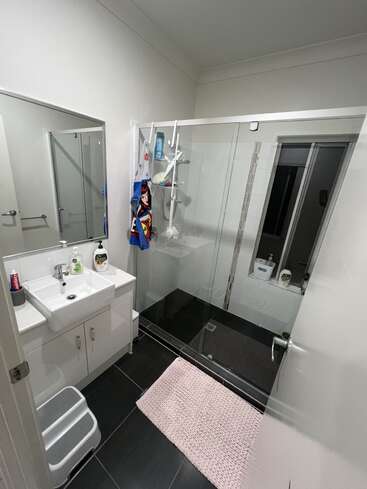 Se trata de un cuarto de baño moderno con ducha de cristal, tocador blanco, alfombra rosa, azulejos negros, espejo, taburete, artículos de tocador, toalla y una ventana.