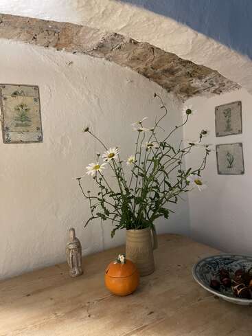The image depicts a rustic wooden table with a vase of daisies, a ceramic pumpkin, and a statue, set against a white wall with decorative tiles.