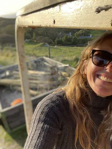 The image shows a woman with long hair and sunglasses, standing in front of a wooden structure, with a blurred background of a field and a house.