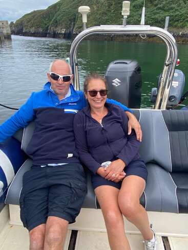 A smiling couple sits closely together on a boat, both wearing dark sunglasses and casual clothes. Green hills and calm water create a scenic background.