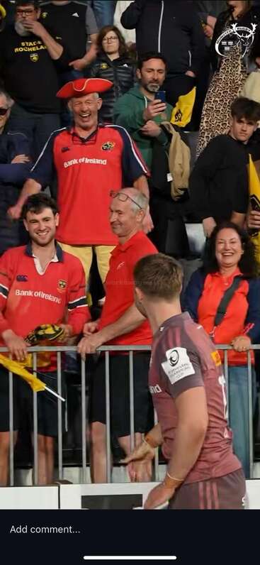 A rugby player interacts with enthusiastic fans dressed in Munster jerseys. The crowd is smiling, cheering, and capturing the moment, with vibrant and lively energy.