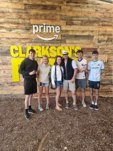 Six people pose and smile in front of a wooden wall with "Prime" and "Clarkson's Farm" signs, likely visiting the site from the TV show.