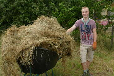 A young man stands outdoors, smiling beside a wheelbarrow overflowing with hay. He wears gloves, casual clothes, and appears to be working in a natural, rural setting.