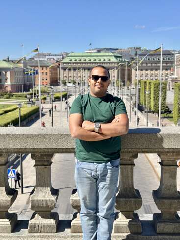Un hombre con gafas de sol y camiseta verde se alza confiado sobre un puente de piedra, con los edificios y banderas de la ciudad de fondo bajo un cielo azul despejado.