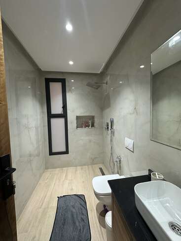 Este moderno cuarto de baño cuenta con paredes de mármol beige, una gran ducha de efecto lluvia, inodoro flotante, lavabo sobre encimera negra, una ventana y lámparas empotradas en el techo. Decoración minimalista.