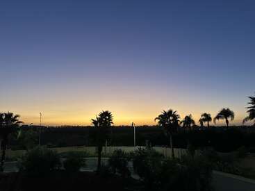 La imagen muestra una tranquila puesta de sol nocturna con un cielo degradado, siluetas de palmeras, farolas y arbustos, creando un ambiente tranquilo y tropical.