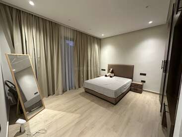 Un dormitorio moderno y minimalista con suelo de madera clara, cama de matrimonio, mesilla de noche, grandes cortinas, un espejo de pared, lámparas de techo y un peluche sobre la cama.