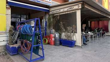 Esta imagen muestra un escaparate en el que se exponen mangueras y tuberías. Al lado, la gente se sienta en las mesas de una cafetería cubierta. Las macetas decoran la acera.