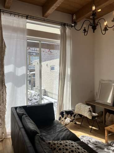 Sunlight fills a cozy living room with wooden beams, a black sofa, a star-patterned blanket, chandelier, cow-shaped decor, sheer curtains, vanity table, and mirror.