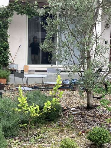 A leafy garden with bushes and trees in the foreground, a small table and chairs, and a person standing behind a window inside the house.