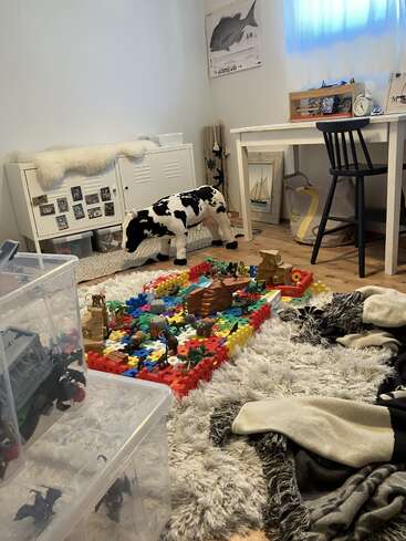 A cozy room with a fluffy rug, scattered blankets, toy storage bins, a table, a chair, a cow plush, and colorful building blocks with animal figurines.