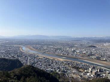 A clear, sunny day view of a sprawling city divided by a winding river with bridges, surrounded by mountains in the distance and urban buildings below.