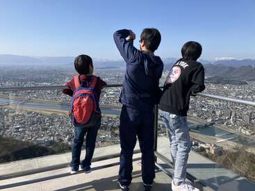 Three people stand at a scenic overlook, gazing at a sprawling city below with mountains in the distance, enjoying the view under a clear blue sky.