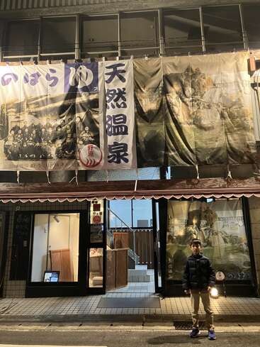 A young boy stands outside a Japanese onsen building at night, illuminated by streetlights. Banners with Japanese characters and historic photos hang above the entrance.